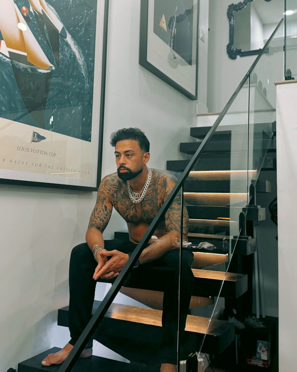 Coach P sitting barefoot on a black staircase, tattooed and wearing silver chains